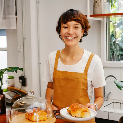 Smiling woman in a brown apron holding a plate with a croissant in a bright kitchen."